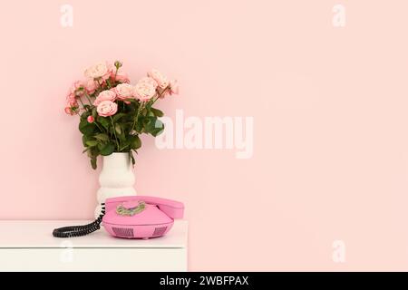 Vase mit Rosenstrauß und Vintage-Telefon auf weißem Schrank in der Nähe der pinkfarbenen Wand. Valentinstag-Feier Stockfoto