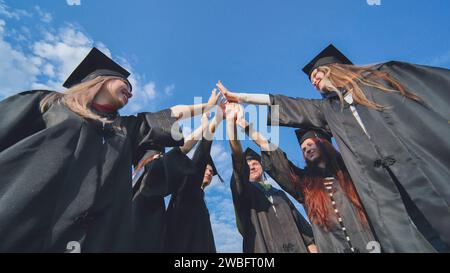 Gruppe glücklicher erfolgreicher Absolventen in akademischen Hüten und Gewändern, die im Kreis stehen und ihre Hände zusammenlegen. Team der Hochschule oder Universität Stockfoto
