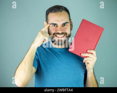 Junger Mann, der mit dem Kopf zeigt, während er ein rotes Buch hält und in die Kamera schaut Stockfoto