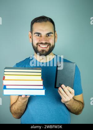 Junger bärtiger Mann mit einem Stapel Bücher in der einen Hand und einem elektronischen Buch in der anderen Stockfoto