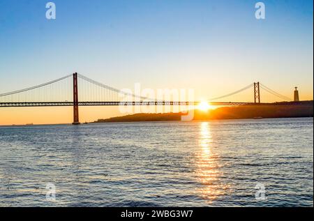 Goldener Sonnenaufgang hinter der Brücke von 25 de Abril in Lissabon, Portugal, der in einem Moment der Ruhe und Schönheit einen warmen Glanz über dem Fluss Tejo ausstrahlt. Stockfoto