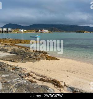 Weißes Ruderboot am weißen Sandstrand von Sommarøy in Troms, Norwegen. Yacht im Hintergrund das türkisfarbene Wasser des Atlantiks mit Inseln Stockfoto