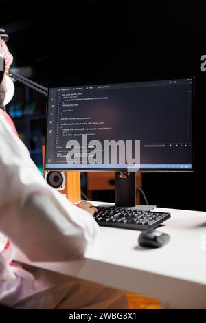 Der muslimische Kerl sitzt am Schreibtisch und kodiert am Computer, um den Entwicklungsprozess der App zu demonstrieren. Überschulteraufnahme eines pc-Monitors mit Laufalgorithmen, die von traditionell gekleideten arabischen Männern produziert wurden. Stockfoto