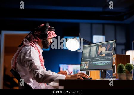 Der arabische Regisseur saß am Tisch und arbeitete aufmerksam an der Filmpostproduktion. Der muslimische Videoeditor in traditioneller Kleidung untersucht Aufnahmen auf dem Computerbildschirm, während er kabellose Kopfhörer trägt. Stockfoto