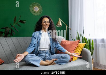 Junge schöne Frau meditiert in Lotuspose, hispanische Frau lächelt zufrieden und blickt in die Kamera, sitzt auf Sofa im Wohnzimmer zu Hause, übt Yoga-Unterricht. Stockfoto