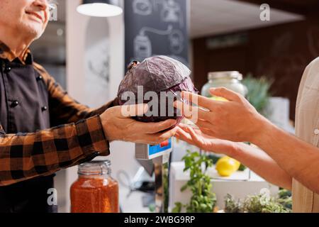 Älterer Mann an der Kasse in der Zero Waste Shop, der Bauernkohl an vegane Kunden mit grünem Lebensstil verkauft. Hipster-Paar kauft Lebensmittel in einem Laden in der Nachbarschaft, Nahaufnahme Stockfoto