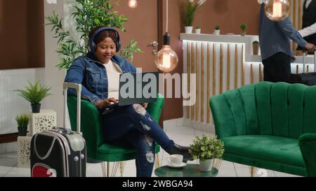 Der Reisende sieht sich eine fernsehsendung im Loungebereich an und wartet auf den Check-in nach einem langen Flug. afroamerikanische Frau mit Laptop in der Lobby des Hotels, pazifische Zeit, bevor sie sich an der Rezeption anmeldet. Stockfoto