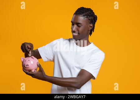 Kluges Sparen. Happy Young Black Guy, Der Eine Münze In Die Sparkasse Einfügt Stockfoto