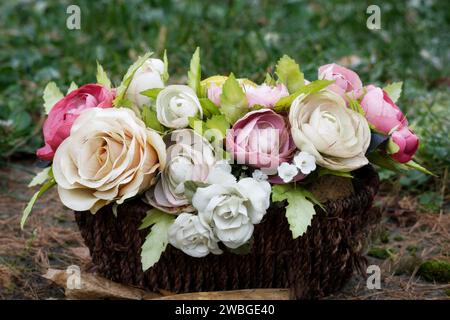 Ein Korb mit pastellfarbenen, verblassten künstlichen Rosen auf einem Grab auf einem Friedhof Stockfoto