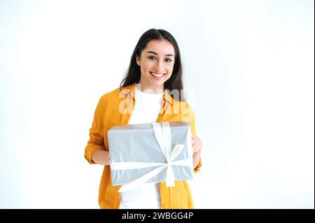 Geschenkkonzept. Fröhliche attraktive brünette inder- oder arabianerin in lässigem orangenem Hemd, steht auf isoliertem weißem Hintergrund, hält große Geschenkbox, blickt glücklich in die Kamera und lächelt freundlich Stockfoto