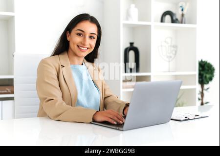 Eine schöne junge, erwachsene indische oder arabische Geschäftsfrau, die Anzug trägt, im Büro sitzt, Laptop bei der Arbeit benutzt, an einem Projekt am Arbeitsplatz arbeitet, die Kamera ansieht und lächelt Stockfoto