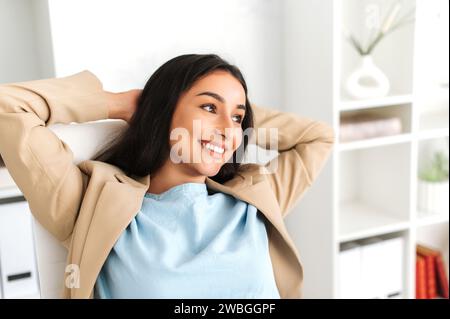 Glückliche zufriedene sorglose Frau indischer oder arabischer Nationalität, die entspannt auf einem Stuhl am Schreibtisch sitzt und ihre Hände hinter dem Kopf hält, wegschaut, eine Pause von der Arbeit einlegt, lächelt, träumt Stockfoto