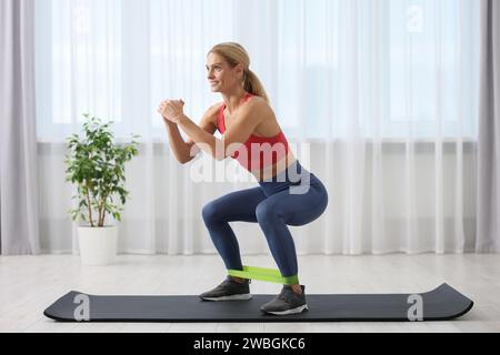 Athletische Frau, die zu Hause Squats mit elastischem Fitness-Band auf der Matte macht Stockfoto