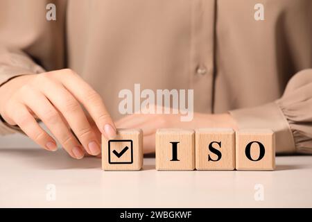 Frau fügt Würfel mit Häkchen zur Abkürzung ISO von Holzwürfeln an weißem Tisch, Großaufnahme Stockfoto