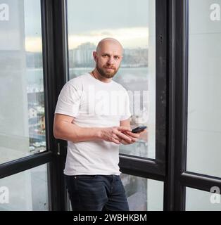Porträt eines jungen Glatzenarbeiters mit Smartphone, während er in einer renovierten Wohnung in der Nähe eines großen Fensters stand. Stockfoto