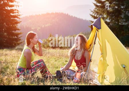 Zwei Wanderfrauen, die in den Bergen campen. Weibliche Freunde, die auf dem Campingplatz Pause machen. Junge, positive Frauen, die auf Gras neben dem Zelt sitzen, lächeln, Tee trinken, reden. Das Konzept der Harmonie mit der Natur. Stockfoto