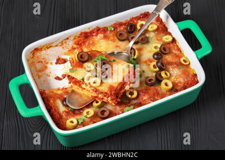 Frisch gebackene Lasagne garniert mit Olivenringen und frischer Petersilie in grüner Auflaufform auf schwarzem Holztisch mit Kuchenschaufel, horizontaler Blick von A Stockfoto