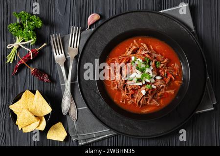 Birria de res, mexikanischer Rindereintopf in heißer Paprikasauce mit roher Zwiebeln und gehacktem Koriander in schwarzer Schüssel auf schwarzem Holztisch mit Gabeln, Taco-Chip Stockfoto