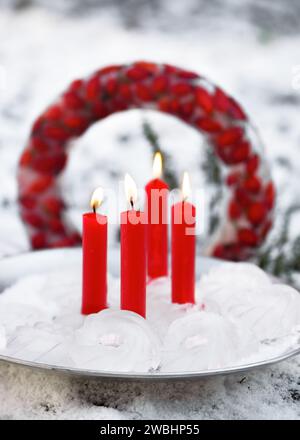 Handgefertigte Garteneisdekoration in Form von Mini-Bundts, mit vier brennenden roten Kerzen auf rustikalem Metallteller vor dem gefrorenen Weihnachtskranz. Stockfoto