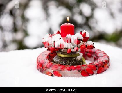 Wunderschöne Blumenarrangements mit Rosenhüftzweigen, gefrorenem Kränz aus Eis, roten Hüftbeeren und brennender roter Kerze im Schnee. Stockfoto