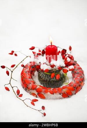 Handgefertigter gefrorener Weihnachtskranz in Form von Ringkuchen aus Eis, rote Rose Hüftbeeren mit brennender roter Kerze im Schnee. Gartendekoration im Freien Stockfoto