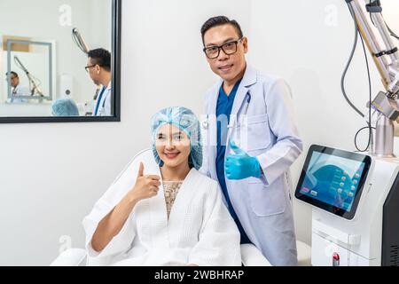 Asiatischer männlicher Kosmetologe und junge Patientin mit blauer Kappe auf dem Kopf sitzend auf Stuhl in der kosmetologischen Klinik, lächelnd, in die Kamera schauend Stockfoto