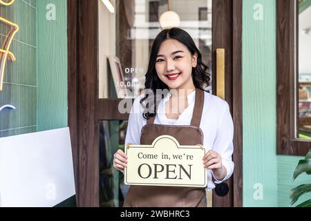 Geschäftsinhaber glückliche, schöne junge asiatische Frau auf Schürze, die in die Kamera schaut, wir sind offen Schild an der Haustür und begrüßen Kunden in einem neuen Café Stockfoto