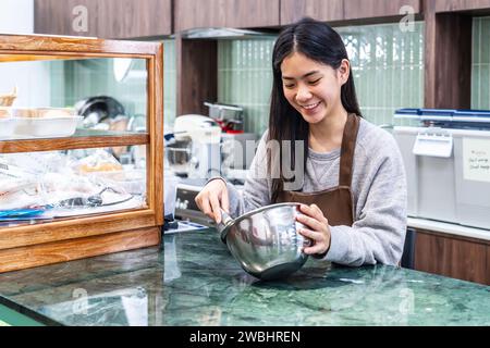 Junge asiatische Frauen sind Ladenbesitzer, ziehen eine Schürze auf, machen Cupcakes-Creme, benutzen einen elektrischen Mixer, bereiten Cookies vor, kochen, hausgemacht, Stockfoto