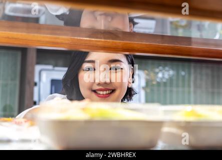 Schöne junge asiatische Arbeiterin, die am Regal stand und frisches Gebäck oder Brot in der Bäckerei des Supermarktes arrangierte Stockfoto