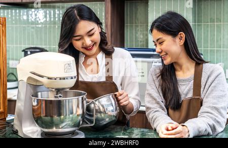 Zwei junge asiatische Freundinnen und Schürzen machen Creme für Cupcakes, mit einem elektrischen Mixer, bereiten Cookies vor, kochen, hausgemacht Stockfoto
