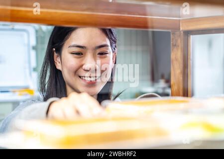 Schöne junge asiatische Arbeiterin, die am Regal stand und frisches Gebäck oder Brot in der Bäckerei des Supermarktes arrangierte Stockfoto
