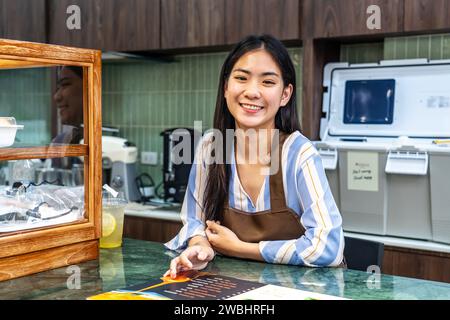 Begrüßung der Gäste, Porträt des fröhlichen jungen asiatischen Profi-Barista oder einer Geschäftsfrau, die eine Bäckerei in brauner Schürze besitzt, begrüßt Gäste in der Nähe der Kassierer Stockfoto