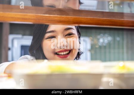 Schöne junge asiatische Arbeiterin, die am Regal stand und frisches Gebäck oder Brot in der Bäckerei des Supermarktes arrangierte Stockfoto