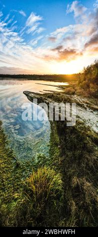 Dieses vertikale Panorama fängt die ruhige Schönheit eines Sees bei Sonnenaufgang ein. Die ersten Sonnenstrahlen küssen den Himmel und malen ihn mit warmen Farbtönen, die sich auf das ruhige Wasser reflektieren. Die Textur des Seeufers, reich an Gras und Algen, führt den Blick auf den fernen Horizont, an dem der Tag beginnt. Die Wolken ziehen sich über den Himmel, spiegeln sich im Wasser unter ihnen und schaffen eine Symphonie aus Farben und Licht, die die stille Majestät des Sonnenaufgangs feiert. Dawn's Reflexion: Gelassenheit am Seeufer. Hochwertige Fotos Stockfoto