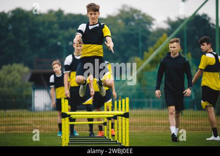 Fußballcamp für Jugendliche. Junge im Teenageralter im Training mit einem Trainer auf dem Grasfeld. Das Sportteam bereitet sich auf die Saison vor. Junge Spieler, die über die Hürde springen Stockfoto