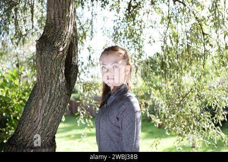 Porträt eines Teenagers, der starken Augenkontakt mit grauem Oberteil unter grünen Blättern des Baumes, Sonnenschein und Baumstamm anstarrt Stockfoto