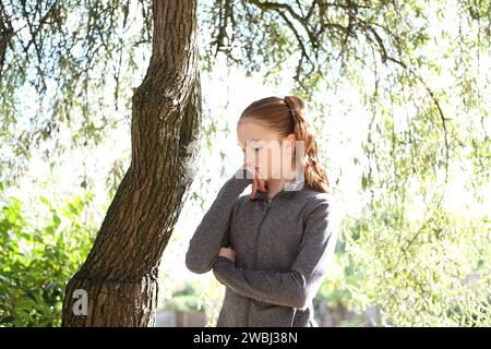 Teenager-Mädchen mit grauem Oberteil unter grünen Blättern von Bäumen, Sonnenschein und Baumstamm, das besorgt und ängstlich aussieht Stockfoto
