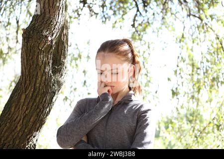 Teenager-Mädchen mit grauem Oberteil unter grünen Blättern, Sonnenschein und Baumstamm, ängstlich und besorgt mit den Augen nach unten Ökoangst Stockfoto