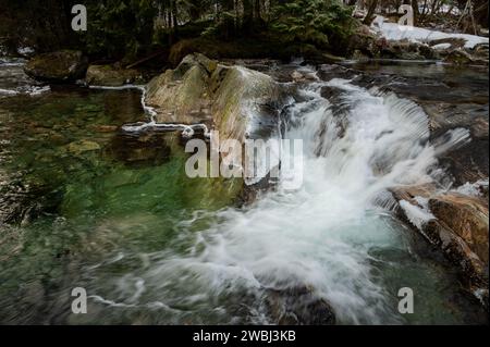 Ein ruhiger Gebirgsbach fließt über moosbedeckte Felsen mit Schneeflächen an den bewaldeten Ufern. Stockfoto