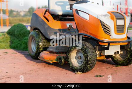 Nahaufnahme des Traktorrasenmähers auf dem Hintergrund eines Haufens gemähten Grases im Park. Rasenpflegekonzept. Stockfoto