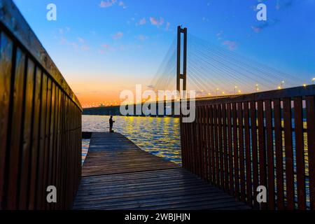 Blick auf einen hölzernen Pier, der zu einem Fischer führt, vor dem Hintergrund einer Kabelbrücke über den Fluss bei Sonnenuntergang. Stockfoto