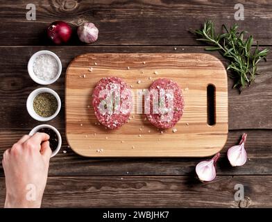 Rohe Fleisch runde Schnitzel - zum Kochen hausgemachter Hamburger. Schnitzel auf einem hölzernen Schneidebrett. Der Koch nimmt Pfeffer zum Würzen des Fleisches. Blick von oben Stockfoto
