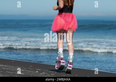Hinten zeigt eine Frau im Sport Kangoo-Jumps-Stiefel, einen schwarzen einteiligen Badeanzug und einen kurzen rosafarbenen Rock, der draußen läuft Stockfoto