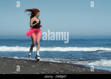 Sportliche Frau im Sport Kangoo springt Stiefel, schwarzer einteiliger Badeanzug, kurzer Rock, Laufen und Springen am Sandstrand Stockfoto