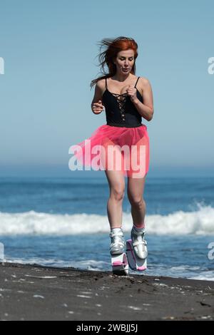 Sportliche Frau im Sport Kangoo springt Stiefel, schwarzer einteiliger Badeanzug und kurzer rosafarbener Rock, der am Strand springt und läuft Stockfoto