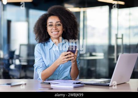 Porträt von jungen Büroangestellten mit Telefon in der Hand, Geschäftsfrau lächelt und in die Kamera blickt, App auf dem Smartphone verwendet, während sie mit Laptop im Büro sitzt. Stockfoto