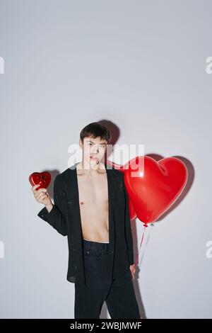 Junger Mann im Hemd mit rotem Herzballon und Geschenk auf grauem Hintergrund, Valentinstag Stockfoto