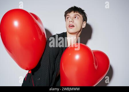 Romantischer Mann in schwarzem Hemd, der rote Herzballons auf grauem Hintergrund hält, Valentinstag Stockfoto