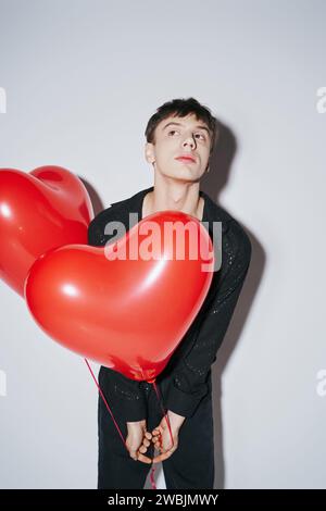 Junger Mann in schwarzem Hemd, der rote Herzballons auf grauem Hintergrund hält, Valentinstag Stockfoto