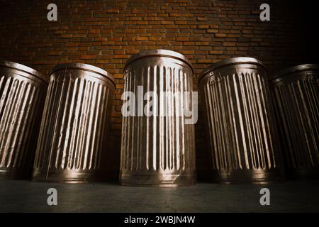 Eine ordentlich angeordnete Formation von glänzenden Metallmüllbehältern, die auf einem Betonboden platziert sind und ihre knackigen Schatten gegen eine alte Ziegelwand in A geworfen haben Stockfoto